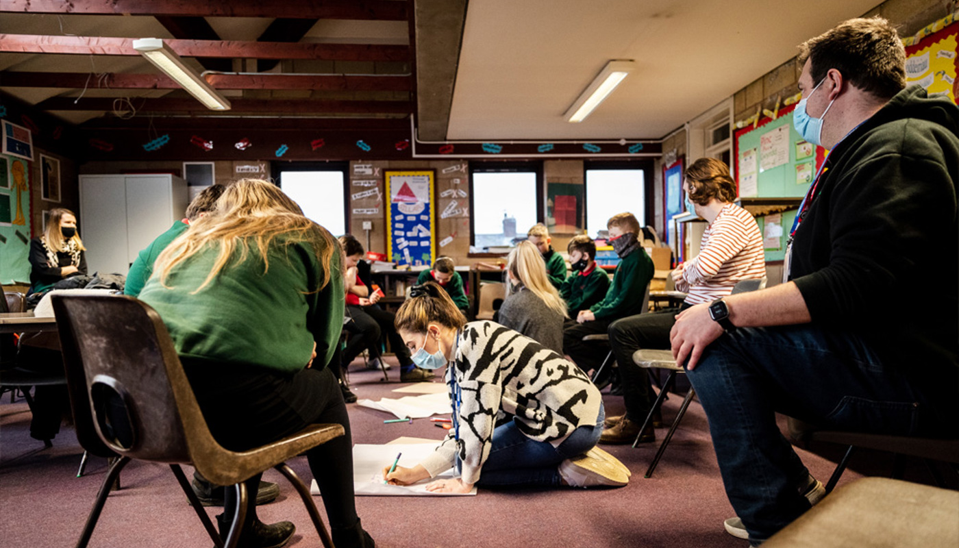 Interior of a busy classroom. A mix of pupils and older individuals are sat on chairs in a circle. One young woman is sat on the floor writing on a large piece of paper and many of the pupils are looking at her. In the background some of the pupils are talking amongst themselves.