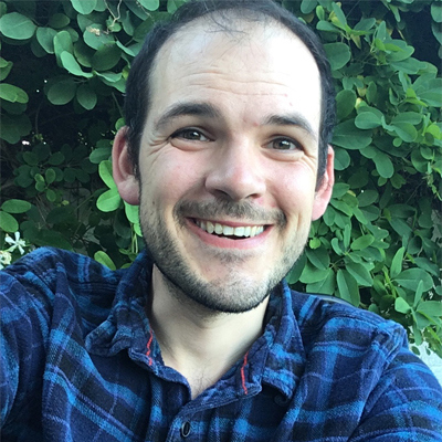 Headshot of Gruffudd Owen. He is a white man with short dark hair and a slight stubble around his chin. He is smiling and looking directly into the camera. He is stood outside in front of green leaves. He wears a blue plaid shirt.