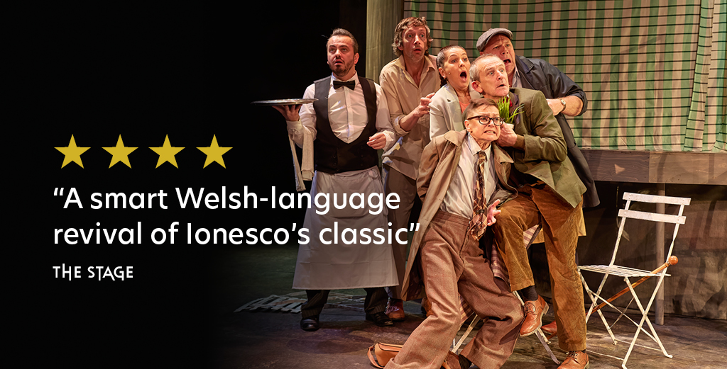 Review graphic, Production image featuring 5 actors onstage, huddled together as though in shock or to protect each other. Overlaid on the image there are 4 small yellow stars. Below this text reads 'A smart Welsh language revival of Ionesco's classic', The Stage
