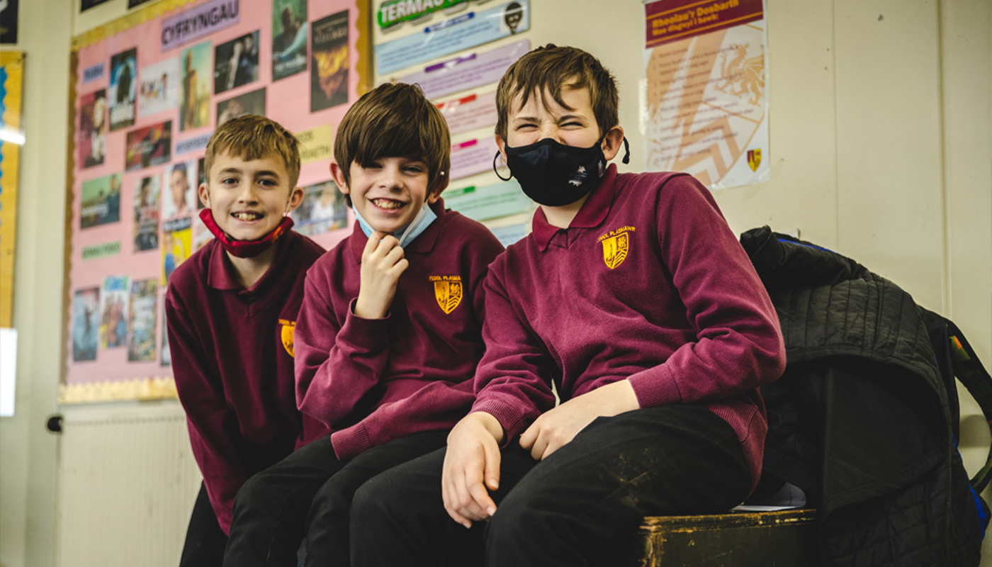 Three young male pupils are sat together on a table. They all wear a school uniform and one is wearing a black face mask. They are all smiling and looking directly into the camera.
