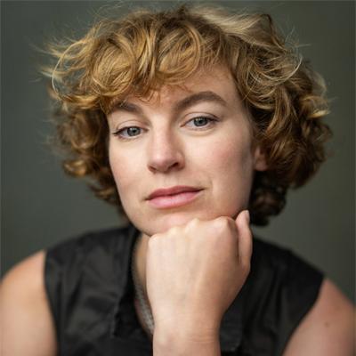 Headshot of Heledd Gwynn. A white woman in her mid-thirties with short mousey coloured curly hair. She is resting her chin on her closed fist, and looking directly into the camera with half a smile on her face.