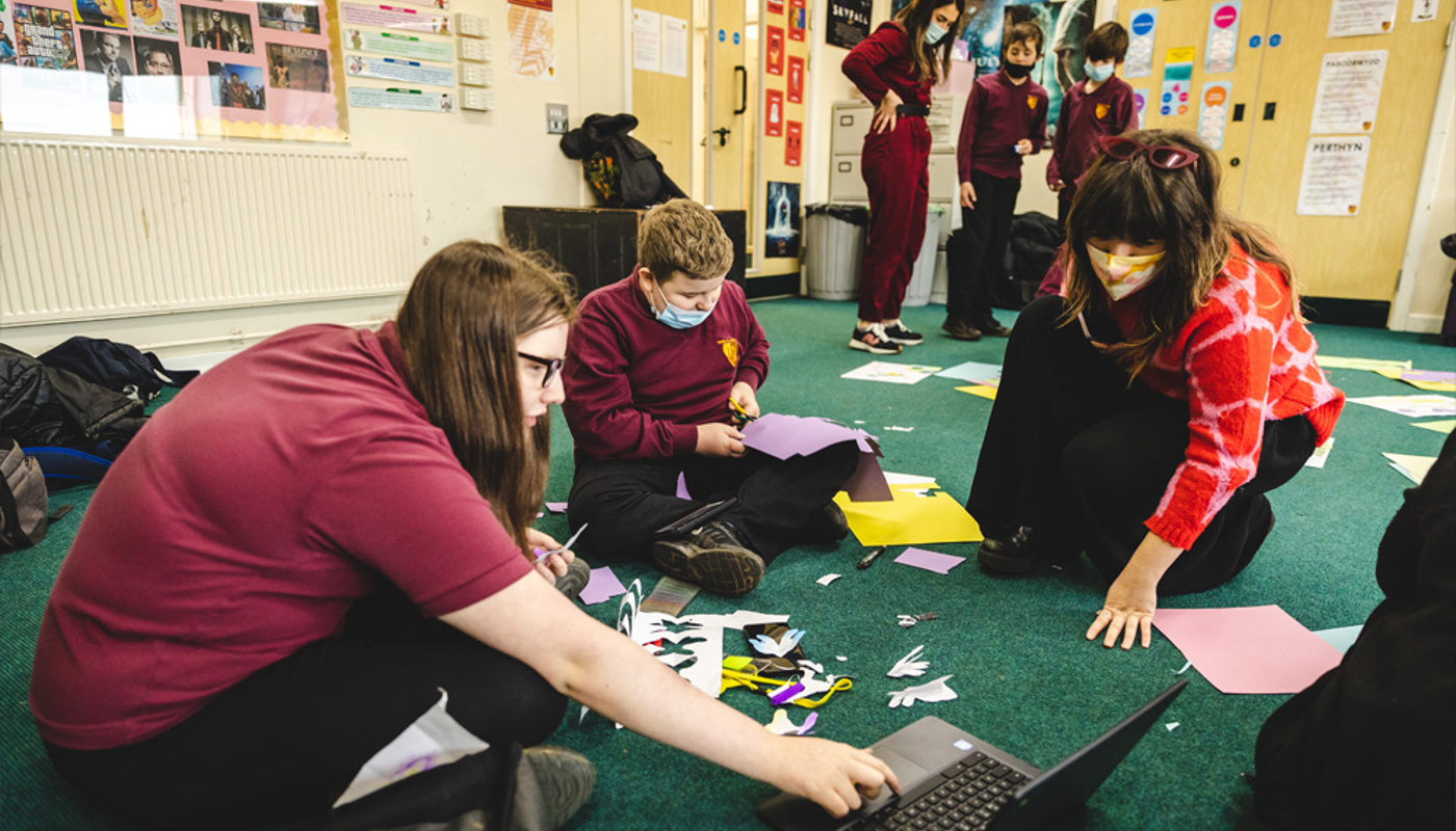 Two young pupils in school uniform and a young woman in formal clothing are sitting on the floor of a classroom. There are pieces of paper and craft supplies on the floor around them. Two are staring down at a piece of work one of the pupils has created, and one is cutting a shape from paper using scissors.
