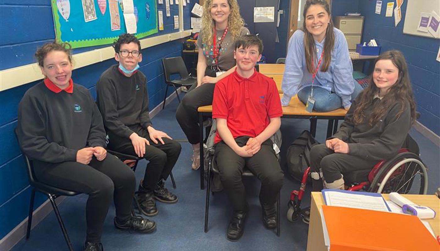 4 young people wearing school uniforms are sat in a classroom. One of them is a young female in a wheelchair. Behind them two young women are sat on top of a table. They wear lanyards around their necks and are dressed formally. Everyone in the image is looking into the camera and smiling. 