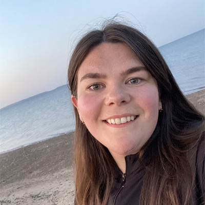 Image of Mared Llywelyn. She is a young, white woman with long dark hair. She is smiling with her teeth and looking directly at the camera. Behind her we can see the beach and the sea.