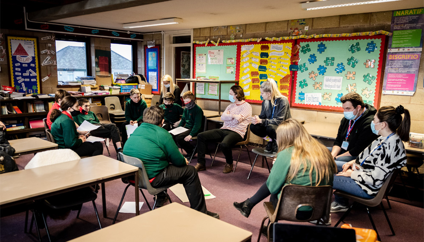 Interior of a busy classroom. The walls are covered in multicoloured informational posters and pupils’ work. A mix of pupils and teachers are sitting in a circle together. Everyone is looking at one of the individuals who is mid conversation. Many of them are wearing face masks.