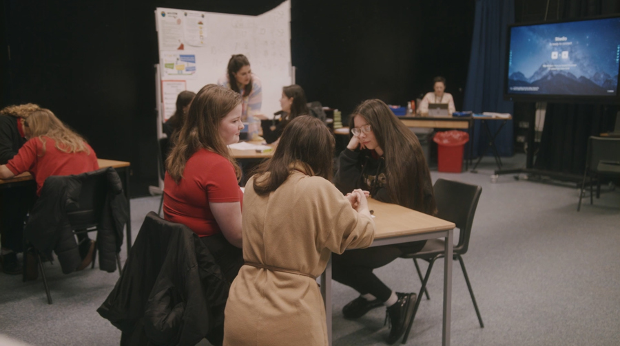 Two young people with dark hair are dressed in school uniform. They are sat opposite each other at a table. A woman kneels down and rests her forearms on the table. She is dressed more formally. Behind them in the background is another group of young people also sat at a table.