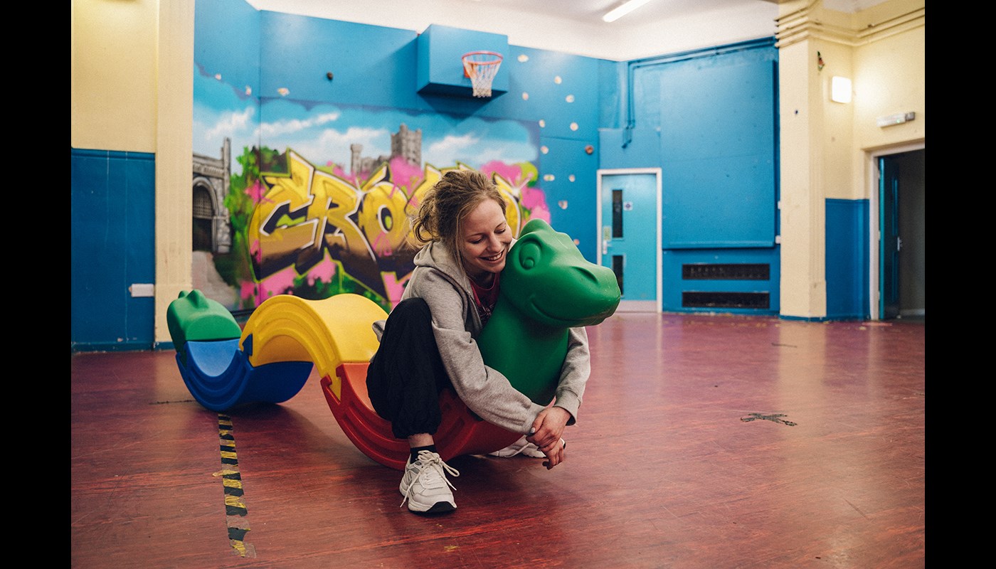 Young woman sits on a green plastic dinosaur with a long colourful tail. She has her arms wrapped around the neck of the dinosaur and looks down smiling.