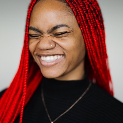 Sarah Adedeji's headshot. Sarah is a black woman with long, red braided hair. She is smiling with her teeth showing and her eyes closed. She is wearing a black top and a small silver necklace with a pendant around her neck. 