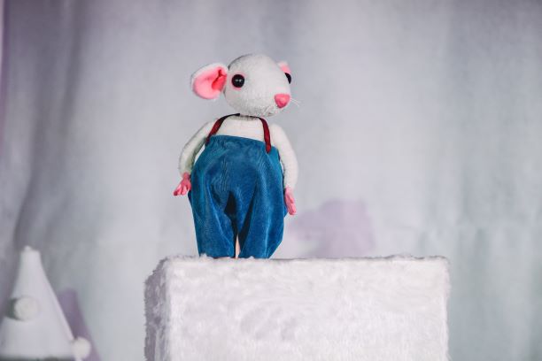 Small plush mouse wears blue corduroy dungarees with tan leather straps. He stands on a textured white block and the background is white. Below him there is a white cone also covered in textured white fabric, with white pom poms stuck to it. 