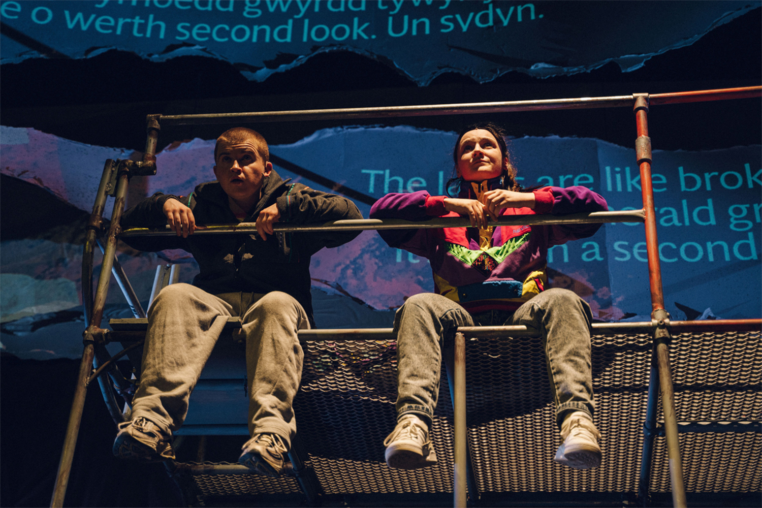 Two young friends sit on top of a climbing frame looking out at the world. Their legs hang over the edge. Both are wearing jeans and trainers. He has a skinhead and wears a drak grey, zip-up jacket. She wears her hair in a ponytail and wears a brightly coloured shellsuit top.