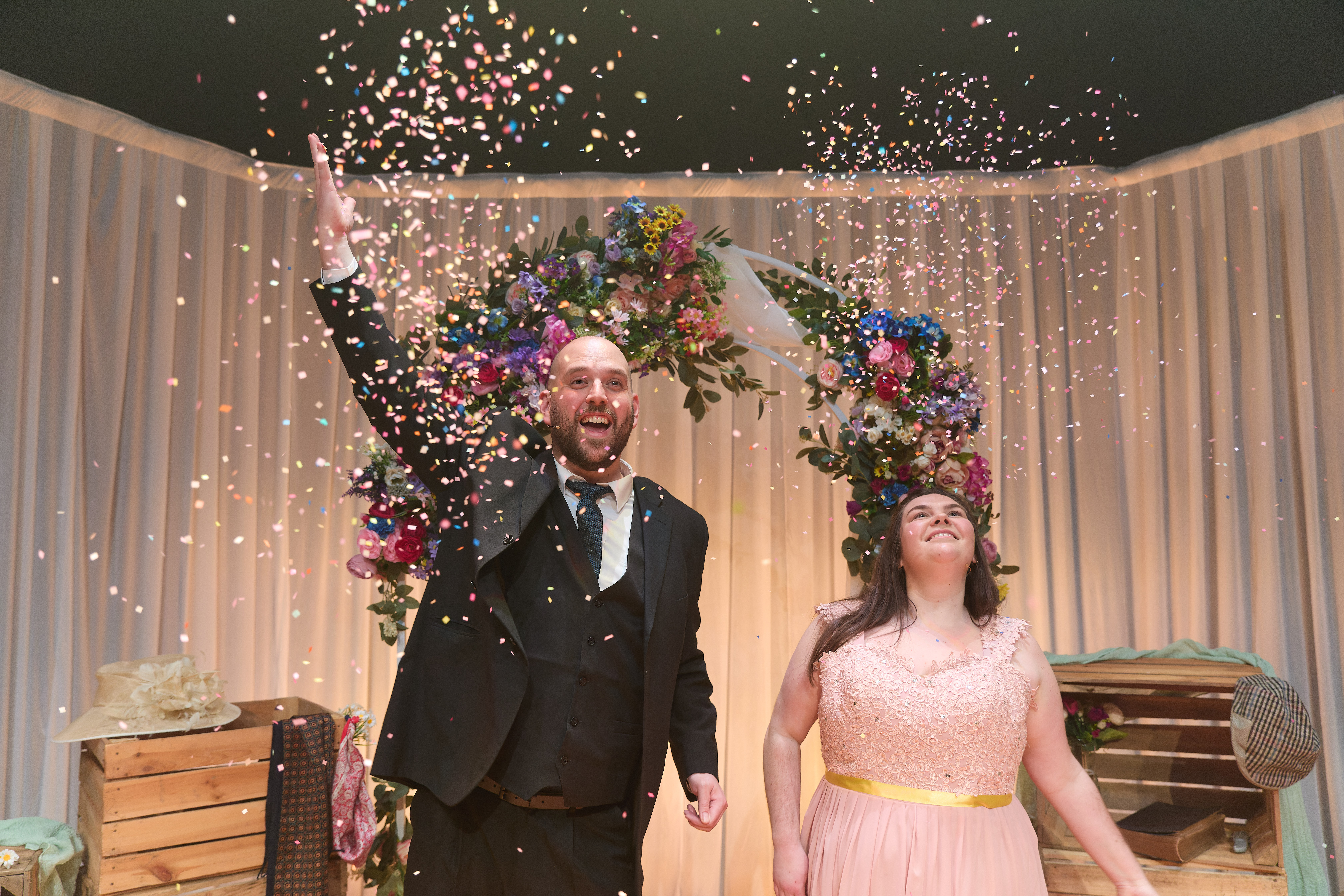Mared Llywelyn and Mark Henry Davies in Parti Priodas. Mared is a young white woman with brown hair. She wears a pink bridesmaid dress. Mark is a young white, bald man wearing a black suit and tie. The set consists of a white drape background, and stacked crates topped with wedding paraphernalia with objects and there is an archway decorated with flowers. Mark is throwing confetti. He smiles and looking at the confetti. Mared is looking up at the confetti with eyes wide, smiling.