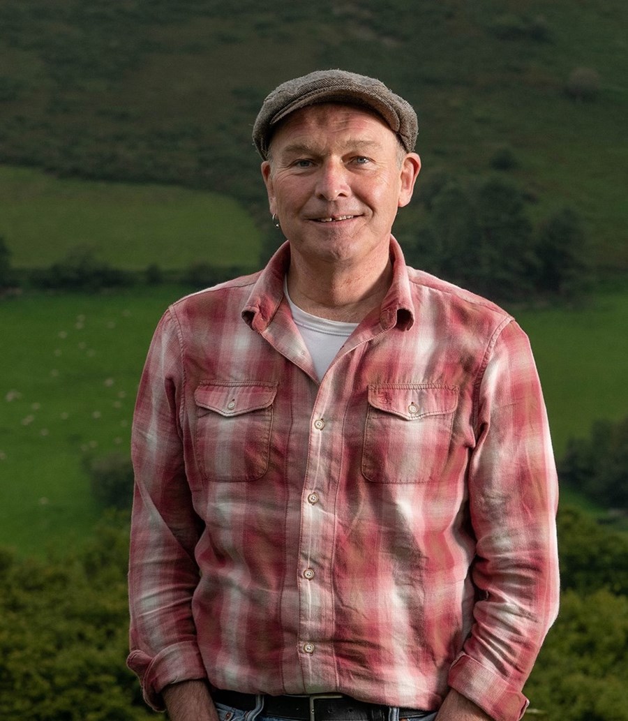 Headshot of Mike Parker. He is stood outside, against a backdrop of green hills. He is a white, middle aged male. He wears a farmer's cap on his head, and has a red plaid shirt on. He is smiling and looking directly into the camera.
