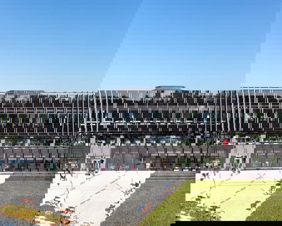 Image of a large office building in the sun. There are hundreds of window panels, and we can see some office furniture through the windows. The sky is blue and there is green grass patches in front of the building. 