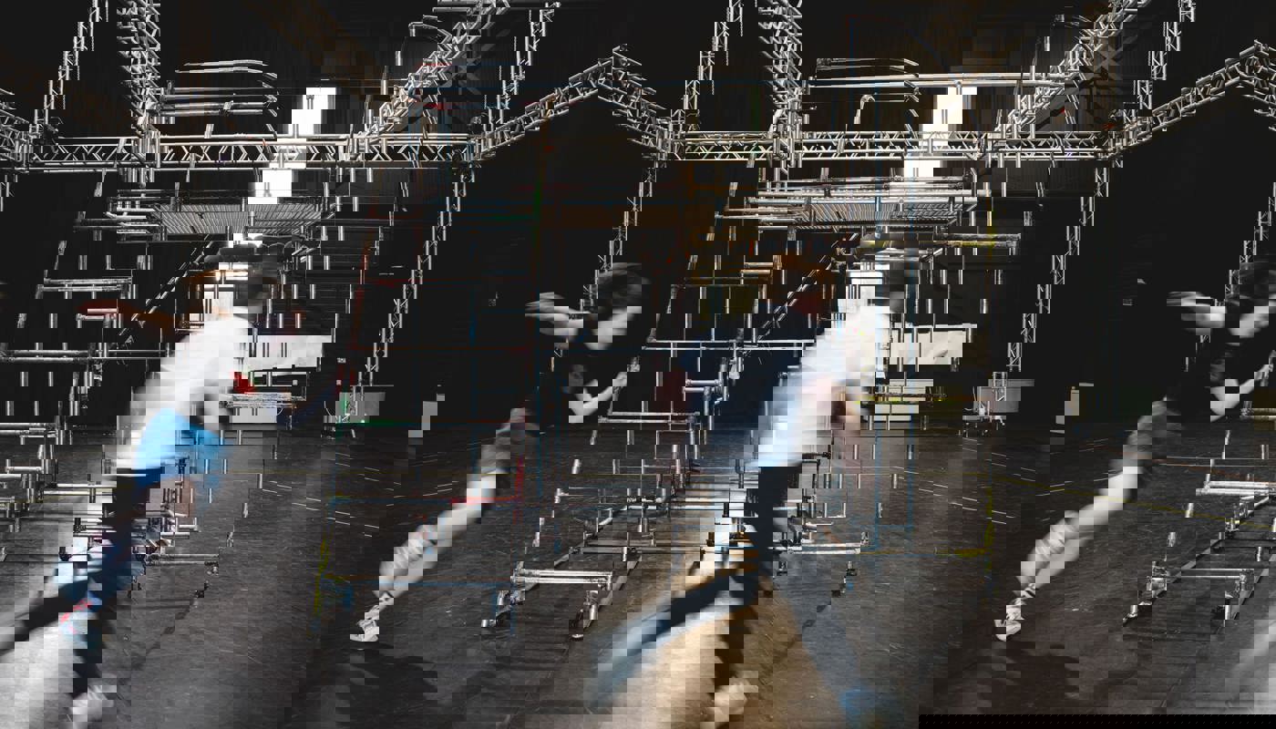 Metal structure built from poles positioned in the middle of a large black room, with metal rigs around the top of the room. Two blurred figures are caught mid-running around the structure.