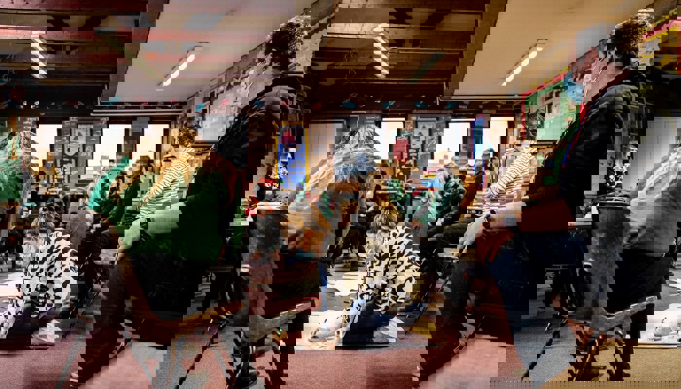 Interior of a busy classroom. A mix of pupils and older individuals are sat on chairs in a circle. One young woman is sat on the floor writing on a large piece of paper and many of the pupils are looking at her. In the background some of the pupils are talking amongst themselves.