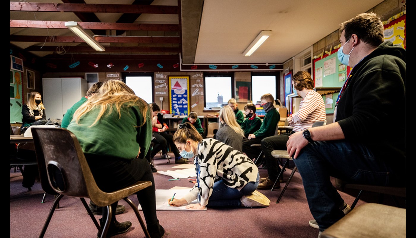 Interior of a busy classroom. A mix of pupils and older individuals are sat on chairs in a circle. One young woman is sat on the floor writing on a large piece of paper and many of the pupils are looking at her. In the background some of the pupils are talking amongst themselves.