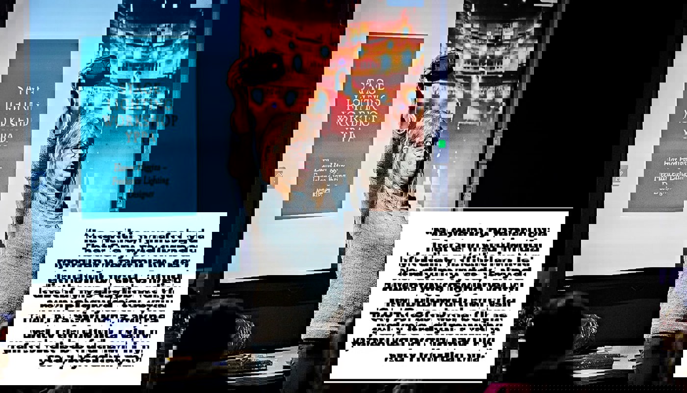 Woman stands in a darkened room in front of a projector screen. Presentation slide on the screen behind her reads 'Stage Lighting Workshop'. She is holding a phone above her face and the torch is on, illuminating her face. She is looking out in front of her, not into the camera. People are sat around her on chairs.