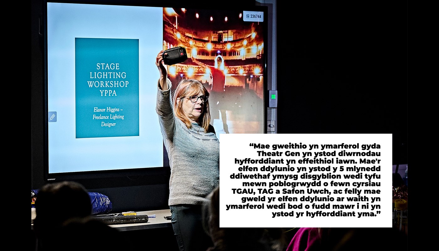 Woman stands in a darkened room in front of a projector screen. Presentation slide on the screen behind her reads 'Stage Lighting Workshop'. She is holding a phone above her face and the torch is on, illuminating her face. She is looking out in front of her, not into the camera. People are sat around her on chairs.