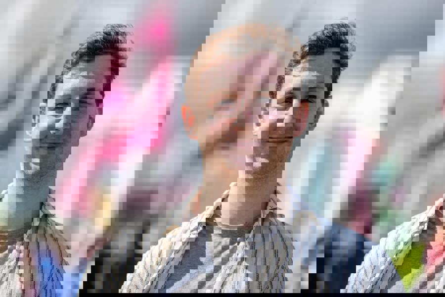 Landscape portrait-style photo of a young man who stands smiling outside. The background is colourful yet blurred.