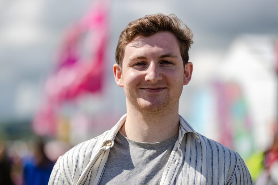 Landscape portrait-style photo of a young man who stands smiling outside. The background is colourful yet blurred.