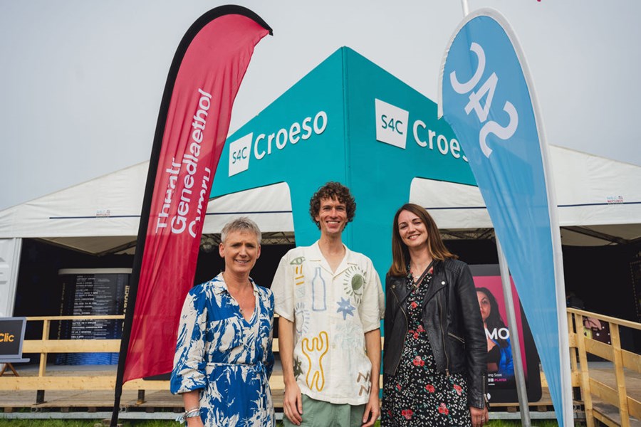 Three people stand in front of banners. The woman on the left has short grey hair and is wearing a blue and white dress. The man in the middle has curly brown hair and an oversized embroidered shirt. The woman on the left has long brown hair and wearing a black leather jacket and floral dress. All three are smiling.