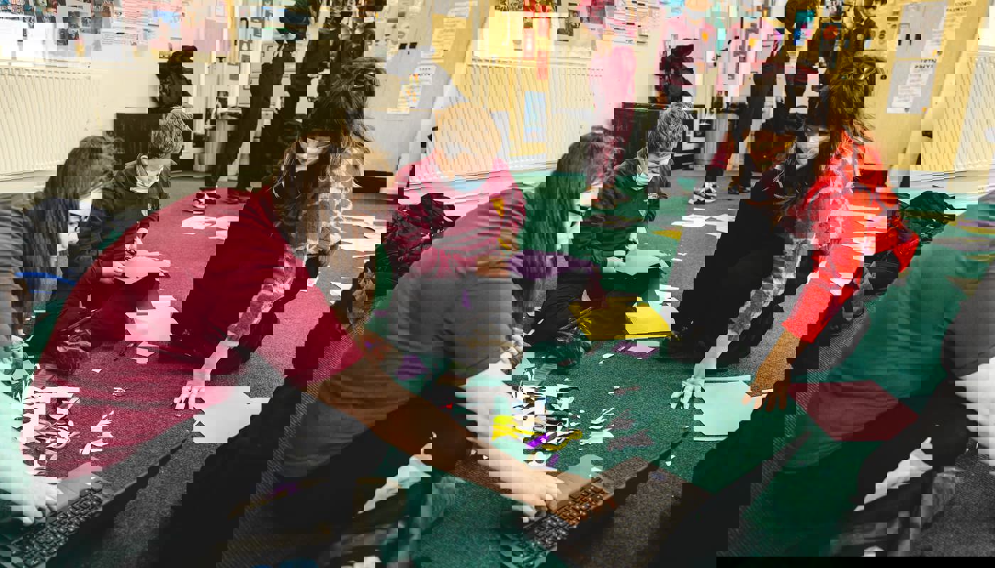 Two young pupils in school uniform and a young woman in formal clothing are sitting on the floor of a classroom. There are pieces of paper and craft supplies on the floor around them. Two are staring down at a piece of work one of the pupils has created, and one is cutting a shape from paper using scissors.