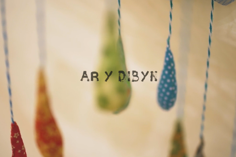 Photograph shows out of focus pieces of multi-coloured fabric dangling on pieces of string. In the centre of the image text reads translated ‘On the edge’