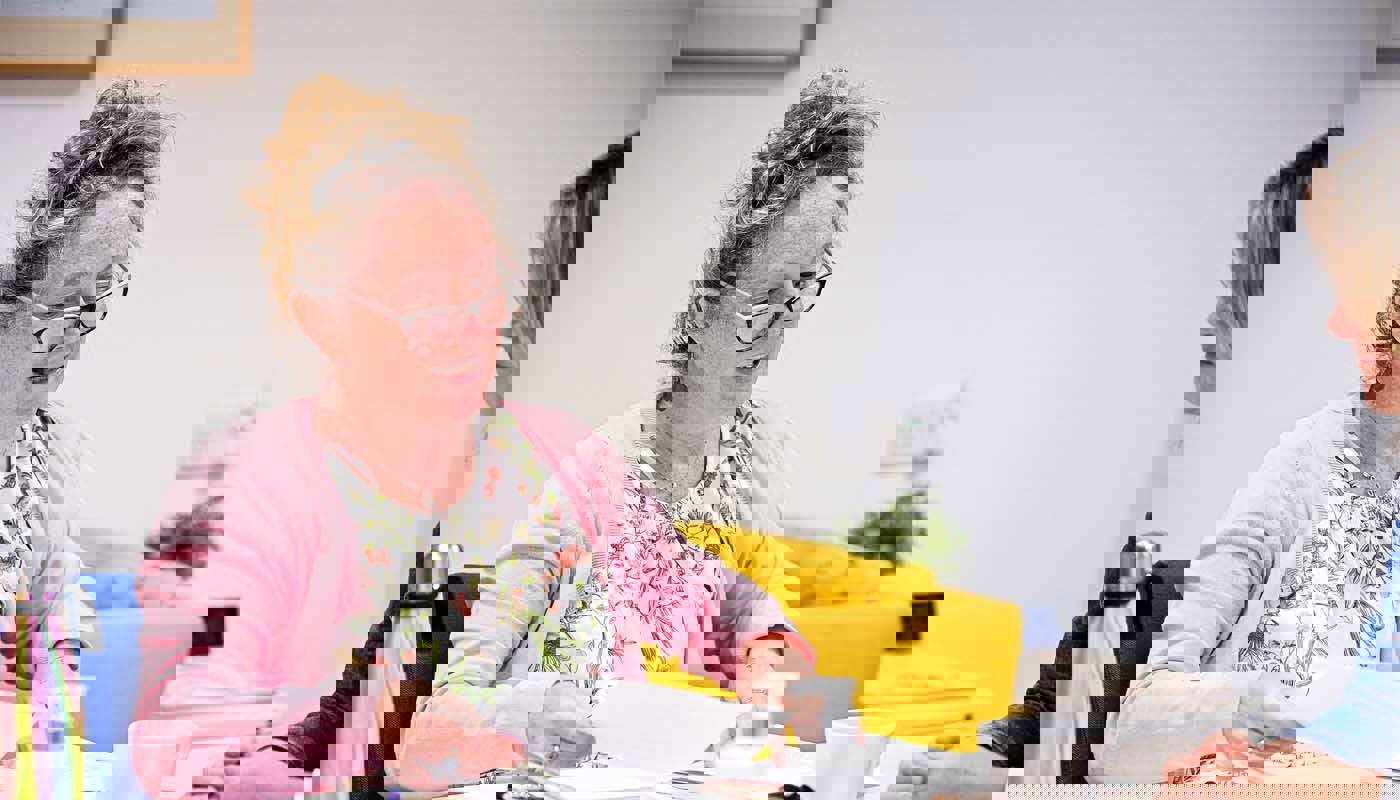 7.	Middle-aged woman sits at a table. Her mouth is half-open. She is holding a piece of paper in her hand and is looking down at it.