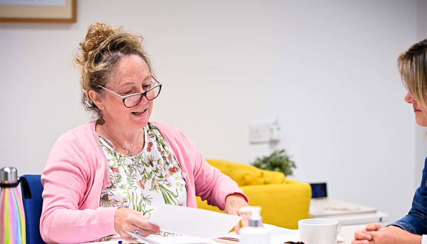 7.	Middle-aged woman sits at a table. Her mouth is half-open. She is holding a piece of paper in her hand and is looking down at it