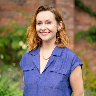 Rhian Blythe's headshot. Rhian is stood outside, there is a blurred brick wall in the background. Rhian is a white female in her thirties. She has strawberry blonde hair reaching her shoulders. She is smiling with her teeth and looking directly into the camera. She is wearing a blue short sleeved shirt and a gold necklace.