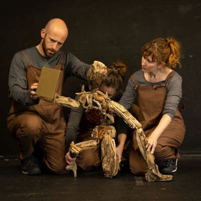 Rehearsal image for Yr Hogyn Pren - three puppeteers dressed in brown and grey are kneeling with a wooden puppet made of drift wood