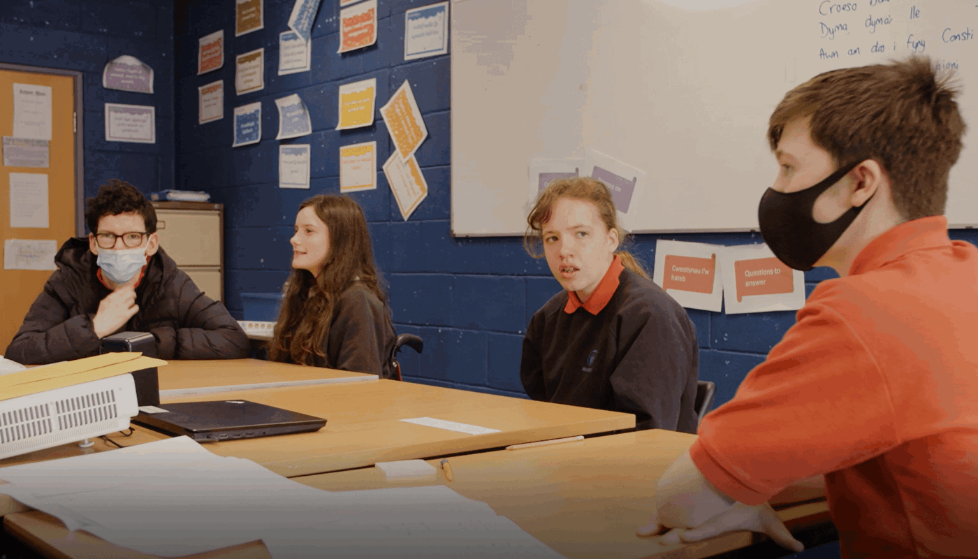 Four young people sit around a table. They all wear a school uniform and Two of them wear face masks. They are all looking at each other. On the table there is a projector and a laptop. There is a whiteboard behind them with unclear blue writing on. On the walls behind them various pieces of paper with colourful writing have been stuck on.
