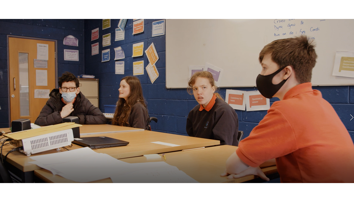Four young people sit around a table. They all wear a school uniform and Two of them wear face masks. They are all looking at each other. On the table there is a projector and a laptop. There is a whiteboard behind them with unclear blue writing on. On the walls behind them various pieces of paper with colourful writing have been stuck on.