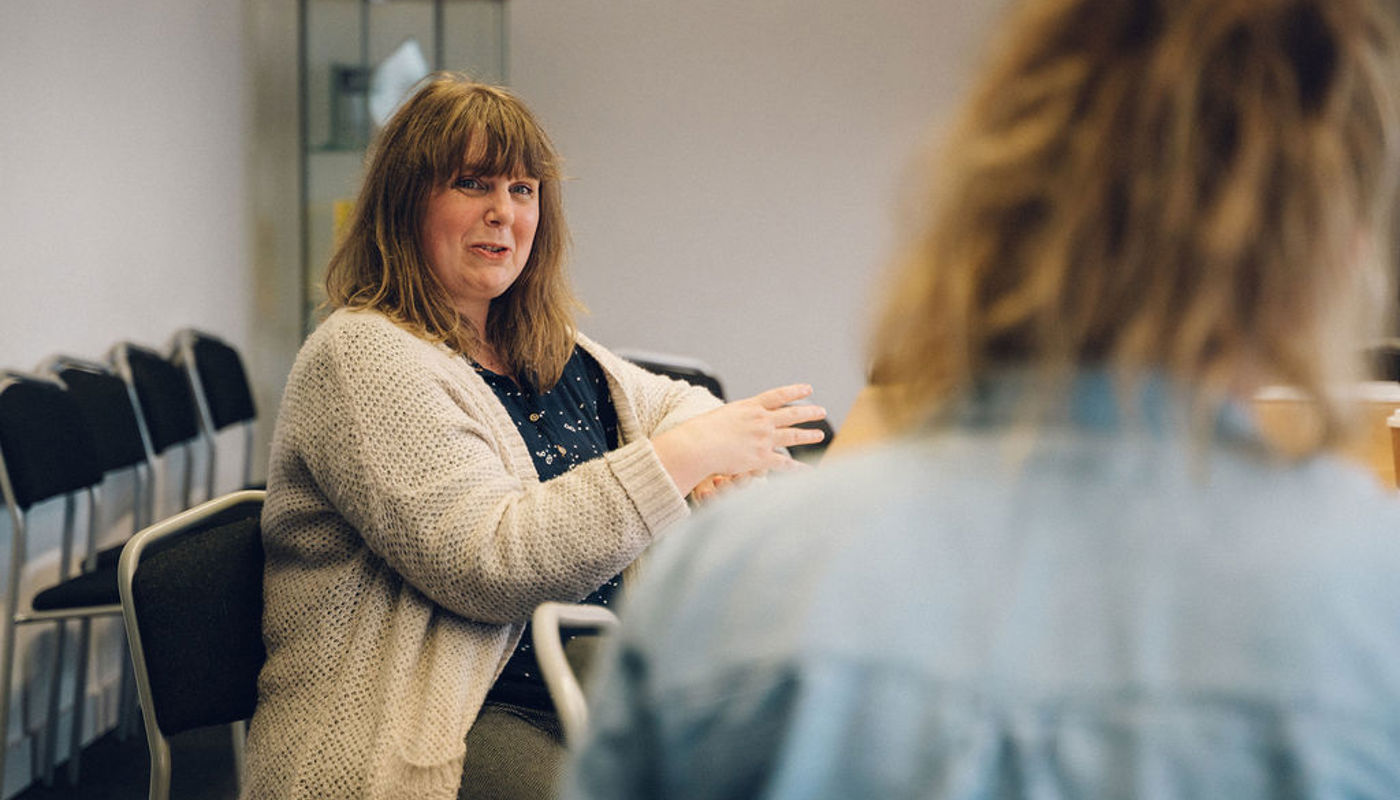 4.	Middle-aged woman sits at a table. Another person sits in front of her with their back to the camera. The woman is looking off camera and has a half-smile on her face. 