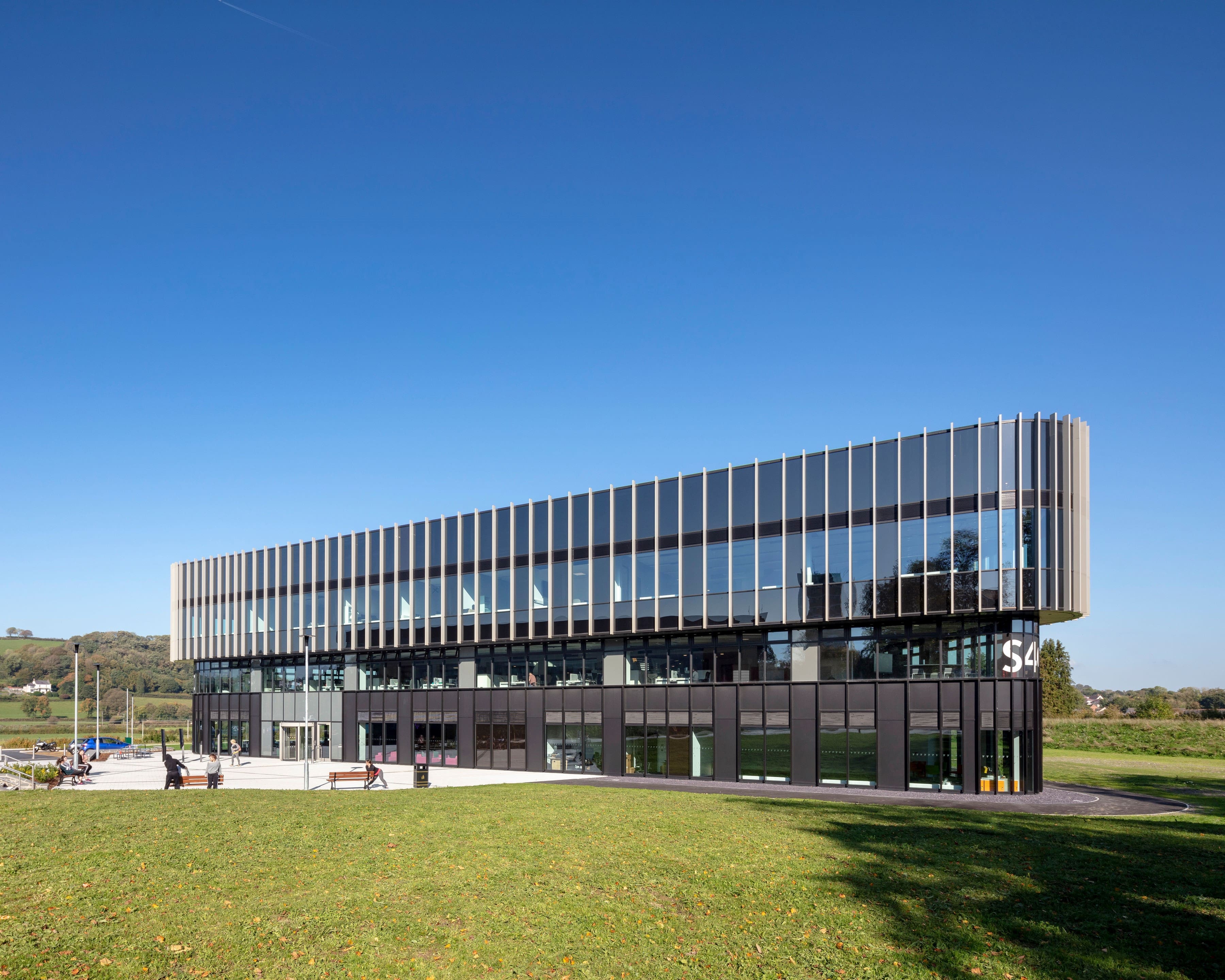 Image of a large office building in the sun from a side-facing angle. It is a very long building, and is not too wide. It has a very modern feel. There are hundreds of window panels, and we can see some office furniture through the windows. The sky is blue and there is green grass patches in front of the building.