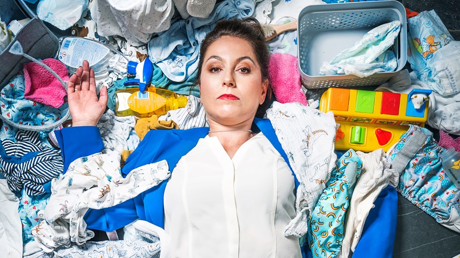 Woman is lying on a pile of baby clothes and other baby necessities like nappies, toys, baby oil and bibs. She is wearing a blue blazer with a white shirt underneath and has red lipstick on her lips. 