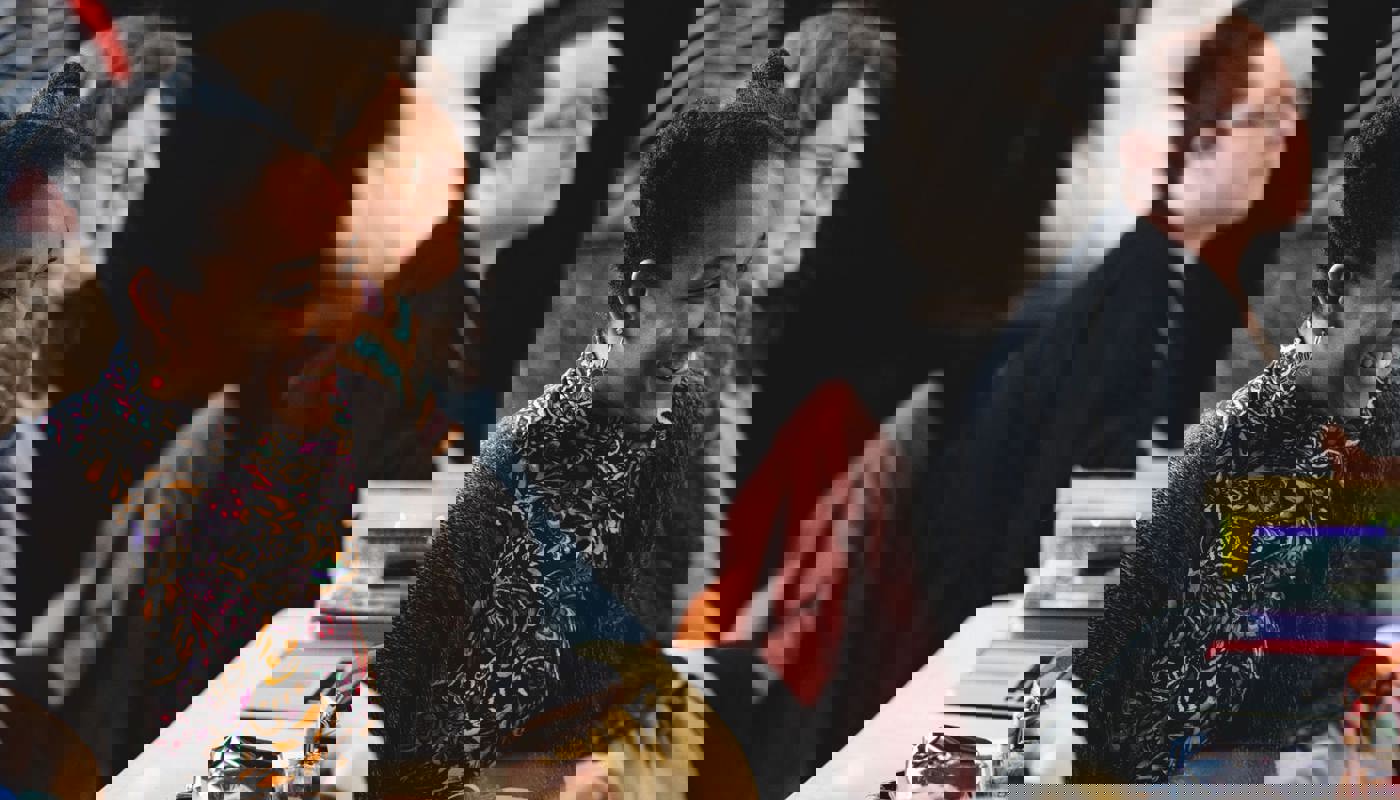 Shot of 4 people sat at a long table. Only one woman is in focus and she is staring out in front of her, away from the camera, and smiling. The other three are not in focus but look ahead in the same direction as her.