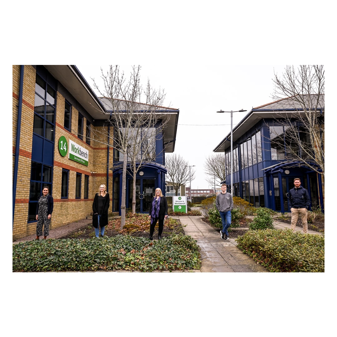 Exterior shot in front of two brick office buildings. There are trees and some bushes, and a concrete path. 5 people stand with distance between each other. There are two older women, one middle-aged woman, a young man and another young man standing. They are all smiling and looking at the camera.