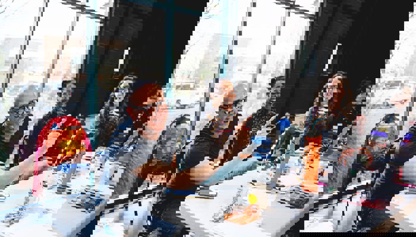 Three people sat at a long table. A man with dark glasses is looking ahead with a smile on his face and pointing a finger in the direction he is looking. A woman is sat next to him smiling with her teeth showing and looks in the same direction as the man. The woman at the end of the table smiles with her teeth showing and looks towards the woman next to her.