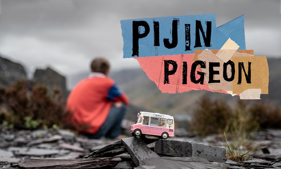 Exterior shot. The sky is grey and there are mountains in the background, unfocused. In front and in focus there is a pink and white small model ice cream van placed on a pile of small slates. Behind it out of focus a boy is sitting down with his back to the camera. Text on image reads ‘Pijin Pigeon’
