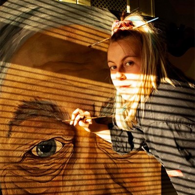 Image of Livia Jones. She is a young white woman with long blonde hair tied up in a ponytail. She is holding a paintbrush in her hand, and she is painting a portrait in front of her. 