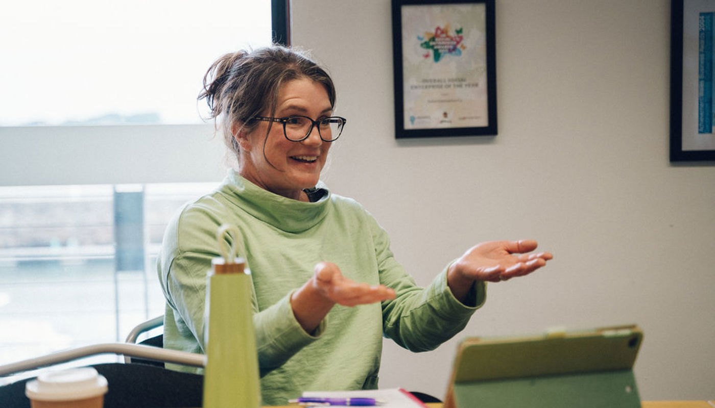 1.	Image of woman sitting down at a table. She is smiling with her mouth open and is holding her arms up mid-way with her palms up. She is looking off-camera. There is a pen, a notebook and an ipad on the table in front of her.