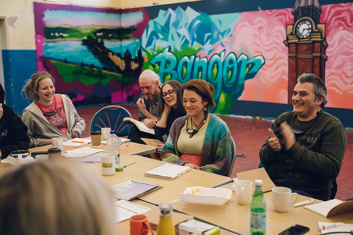Image of people of varying ages sat at a table together. Almost everyone is smiling and laughing. There are pieces of paper, pens and water bottles all over the table. There is graffiti on the wall behind them with the words 'Bangor' painted in the middle.