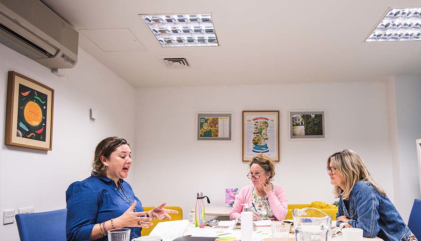 9.	Two middle-aged women and a younger woman sit around a table. The young woman has her mouth open, her hands are up and she is looking at a piece of paper on the table in front of her. The two other women are looking at the young woman.