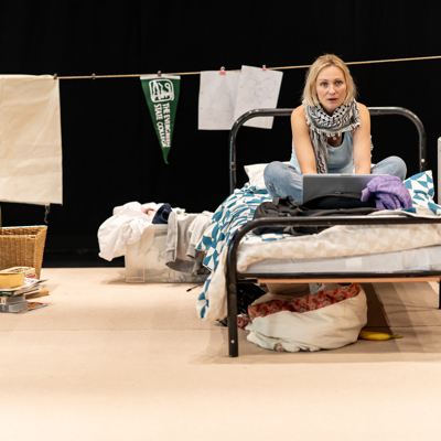 Rehearsal photo for Fy Enw i yw Rachel Corrie - a blonde white woman sits on an untidy bed with an open laptop in front of her. She's wearing a keffiyeh scarf.