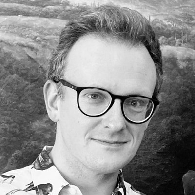 Black and white image of Elidir Jones. He is a man in his thirties. He has short, dark hair and wears black rimmed glasses. He is slightly smiling and looking into the camera.