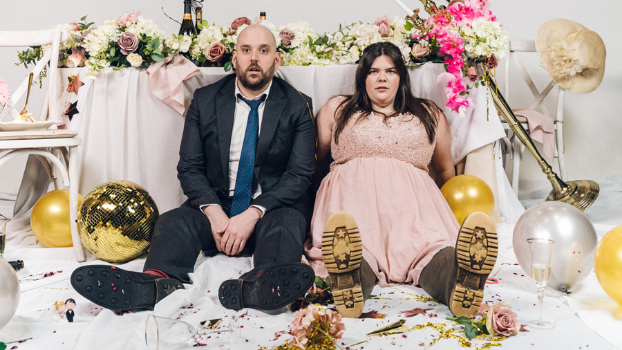 Poster image of Mared Llywelyn and Mark Henry for parti priodas. They are dressed formally in a suit and bridesmaid dress, but Mared has muddy wellingtons on. Around them, balloons, confetti, roses and other party decorations are strewn. Both look directly in front of them and neither are smiling