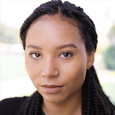 Mali Ann Rees's headshot. She is a young black woman. She has long dark hair worn in braids. She has brown eyes and is looking directly into the camera. She is smiling very lightly.