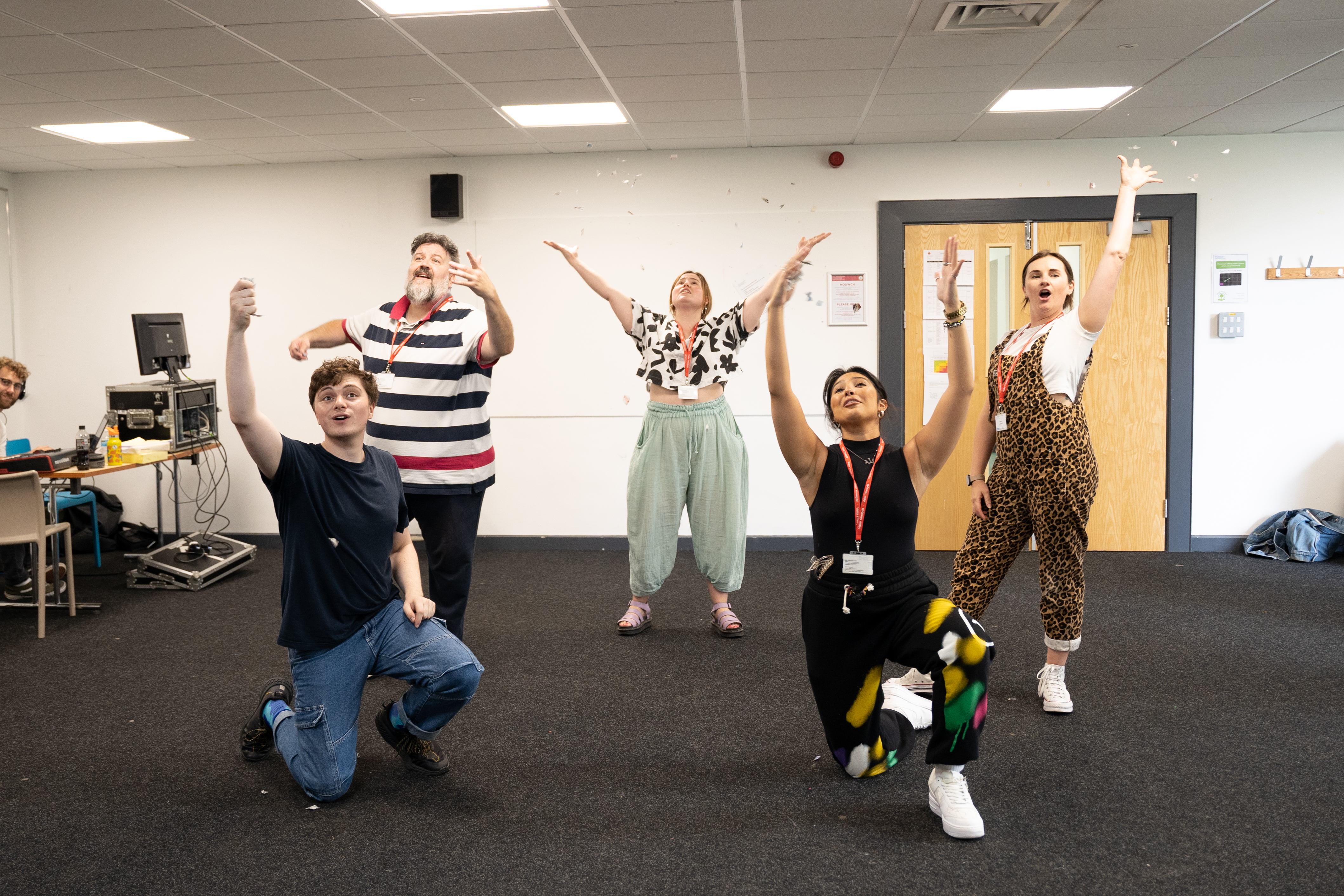 Image of 5 actors in rehearsals for Rwan Nawr. The same actors are returning for Ha/Ha. From left to right: Dewi Wykes is a white man in his twenties kneeling on the ground. Behind him Dion Davies stands, a white middle-aged man with arms outstretched. Caitlin Drake stands in the middle, a young white woman with arms stretched to the sky. In front of Caitlin is Leilah Hughes, a young mixed-race woman kneeling with arms stretched to the sky. Behind Leilah is Lowri Gwynne, a white woman in her thirties. .