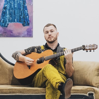 Image of Sam Humphreys. He is a white man in his early thirties. He has very short mousy coloured hair. He is wearing distressed yellow and black dungarees with a dark tshirt underneath. He is sat down on a sofa and is holding an acoustic guitar; his left hand is on the frets, and his right hand is holding a plectrum and appears to be strumming. 